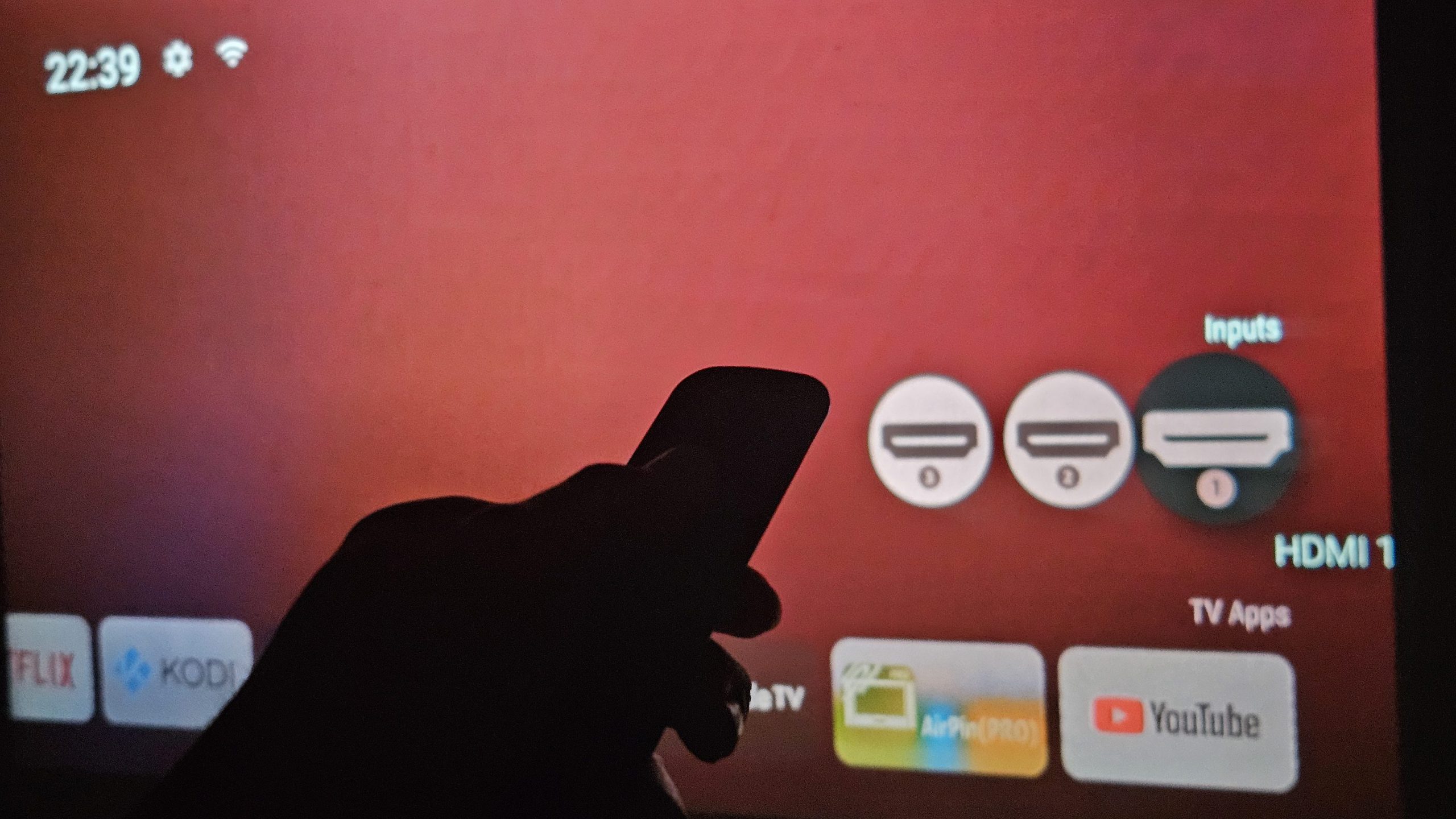 התמונה מציגה יד שחורה גדולה ומטושטשת מצביעה או בוחרת אפליקציה על ממשק חוויה אדום של מקרן HY320 ביתי. ניתן לראות סמלי אפליקציות מוכרות כמו נטפליקס, קודי, YouTube ועוד. היד הגדולה מרמזת על בחירה או שימוש בתוכן ובשירותי מדיה דיגיטליים. הצבעים האדומים והשחורים מעניקים מראה עז וצבעוני לממשק המסך.