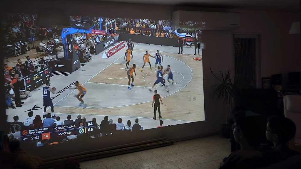 התמונה מציגה רגע בו אנשים צופים במשחק כדורסל, שמוקרן בגודל 155 אינץ' על קיר גדול בבית. 