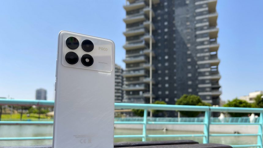 הסמארטפון POCO F6 Pro בצבע לבן על רקע אגם מלאכותי ובניין רב קומות.